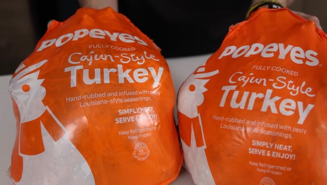 Two orange Popeyes bags labeled "Cajun-Style Turkey" sit on a white surface. The packaging states the turkeys are fully cooked, hand-rubbed with Louisiana-style seasonings, and ready to heat and serve.