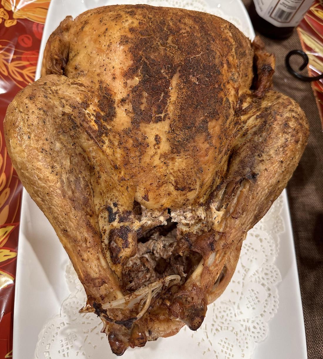 A whole roasted turkey with crispy, browned skin sits on a white platter with a decorative paper doily beneath it, placed on a festive tablecloth with red and orange leafy patterns.