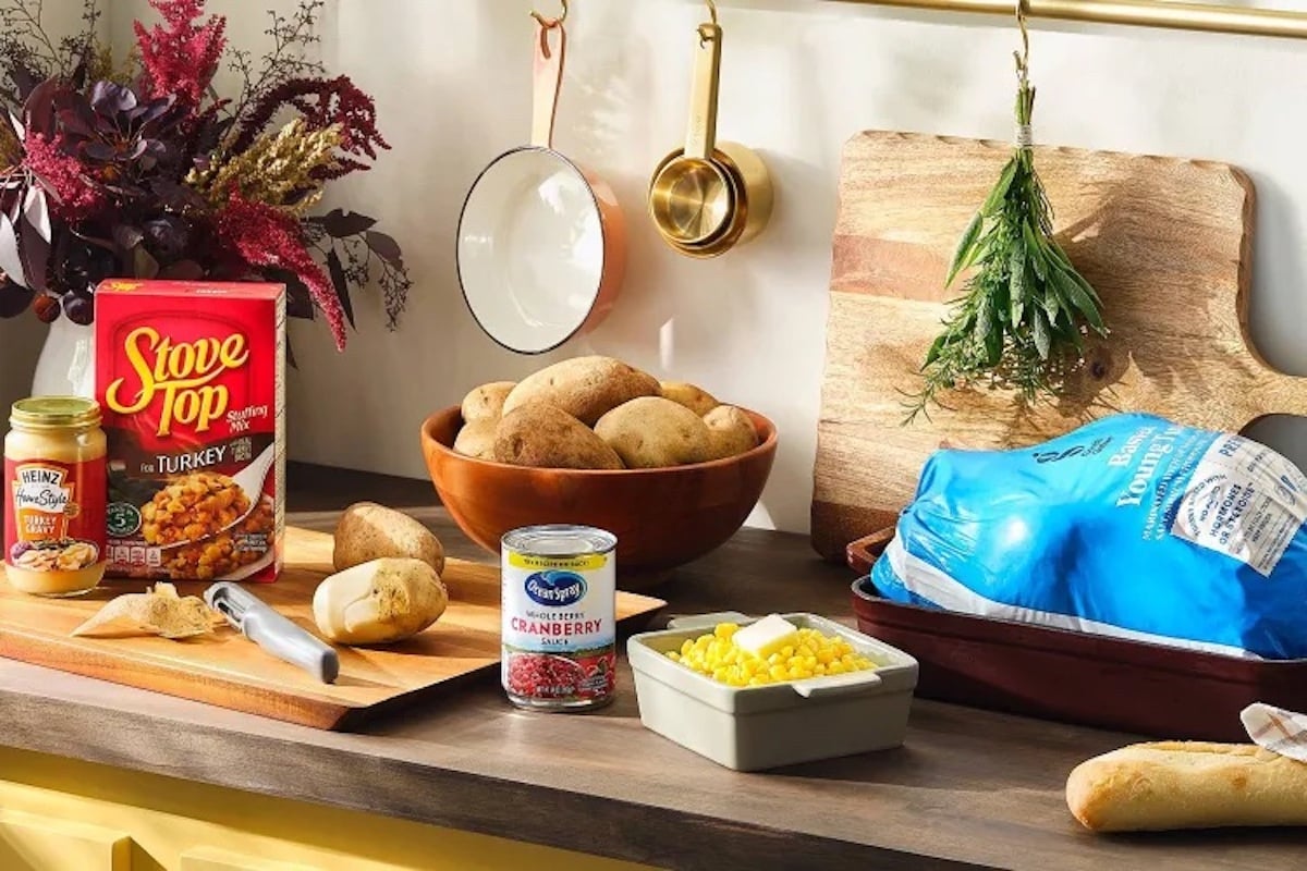 A kitchen counter with a bowl of potatoes, a frozen turkey, stuffing mix, canned cranberry sauce, canned corn, rolls, a cutting board with herbs, and a vase with flowers. Cooking utensils hang on the wall.