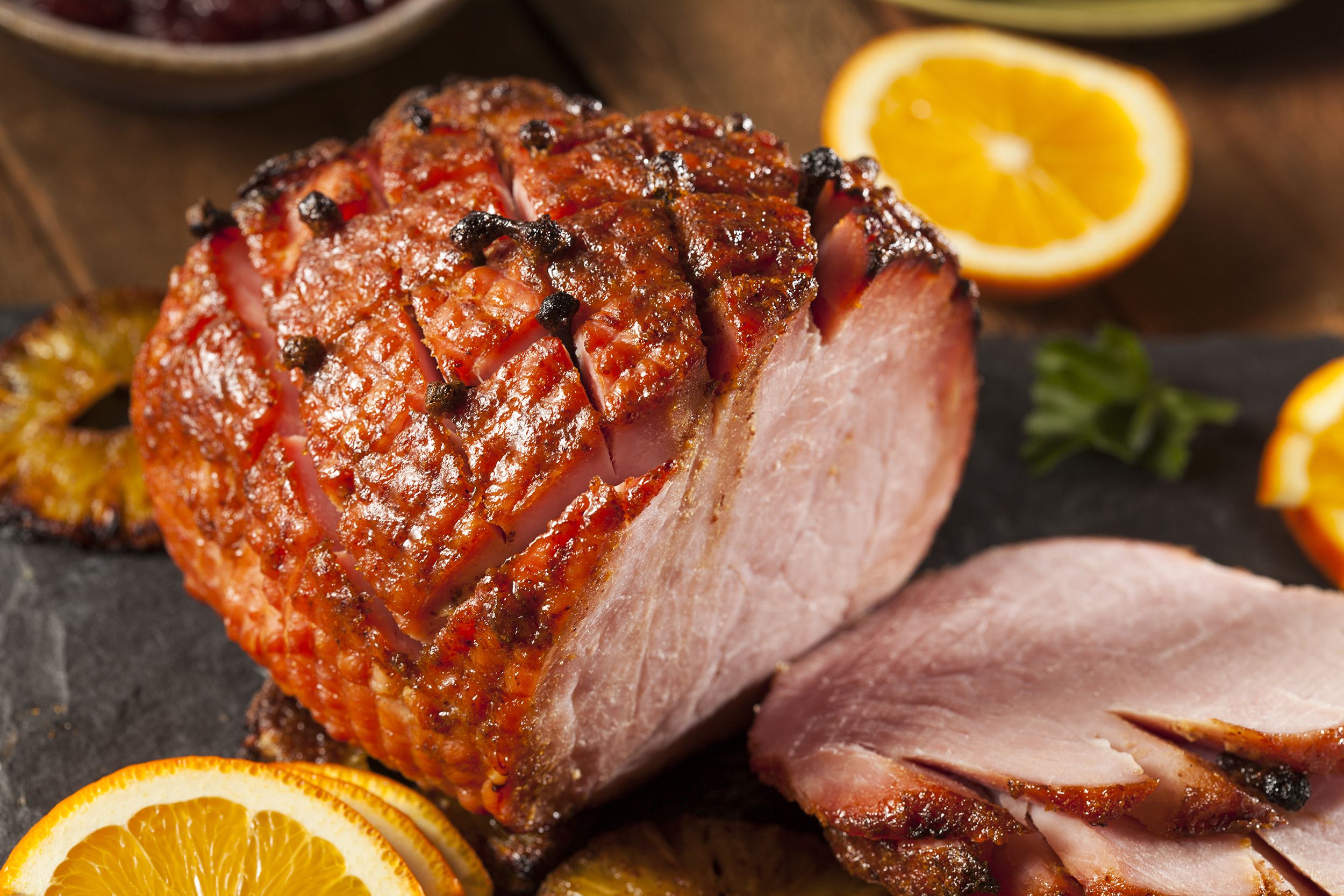 Closeup of baked ham (with brown sugar glaze) with slices surrounded by orange and pineapple slices with a blurred background