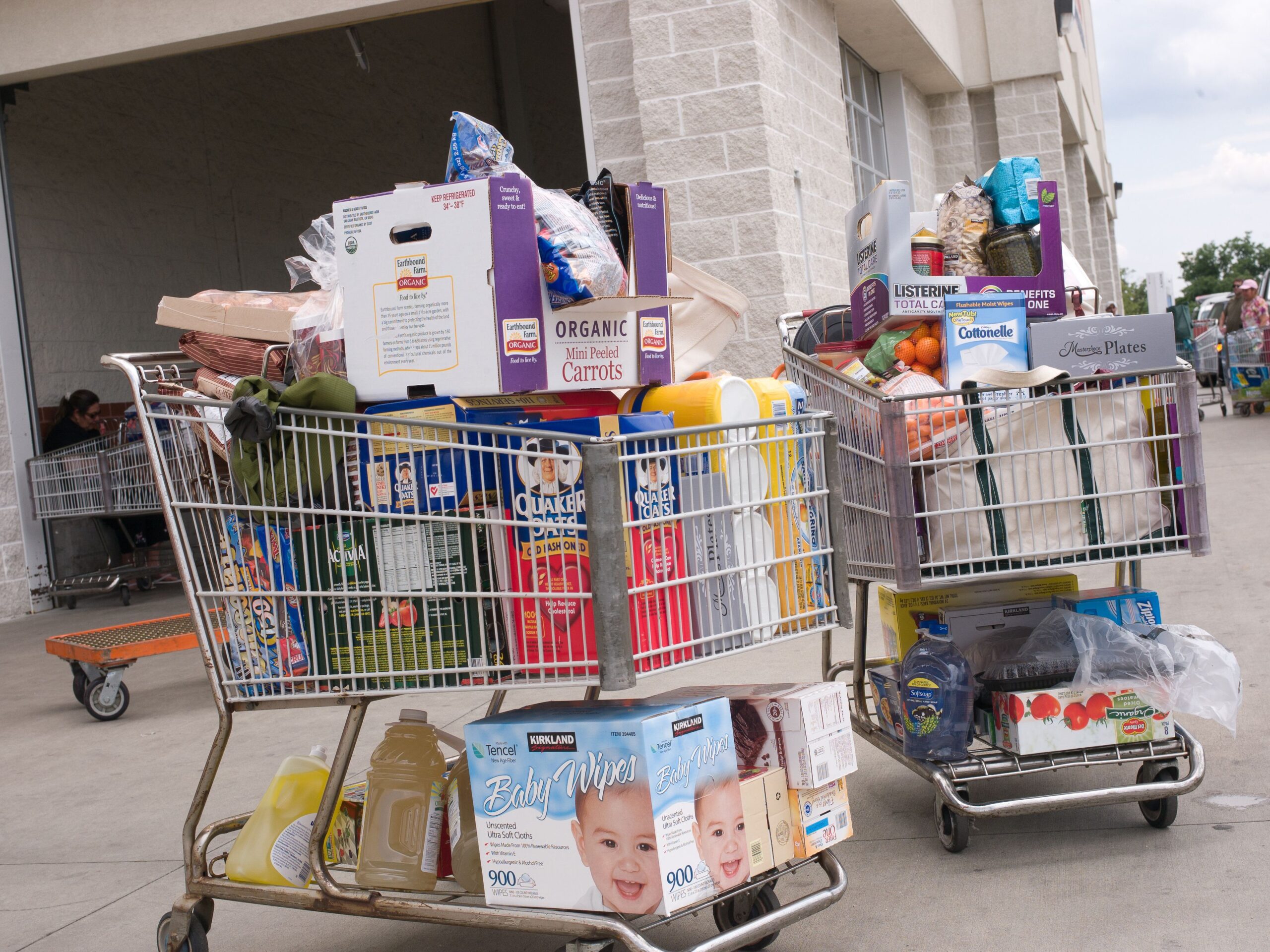 Two shopping carts filled with groceries and household items, including baby wipes, cereal, carrots, oranges, bottled water, and other assorted products, outside a store entrance.