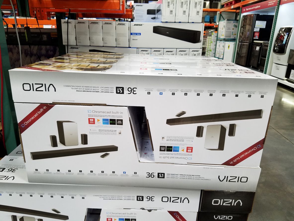 Stacked boxes of Vizio sound bar systems with subwoofers and speakers are displayed on a store shelf. The packaging highlights features like Chromecast built-in and 36\