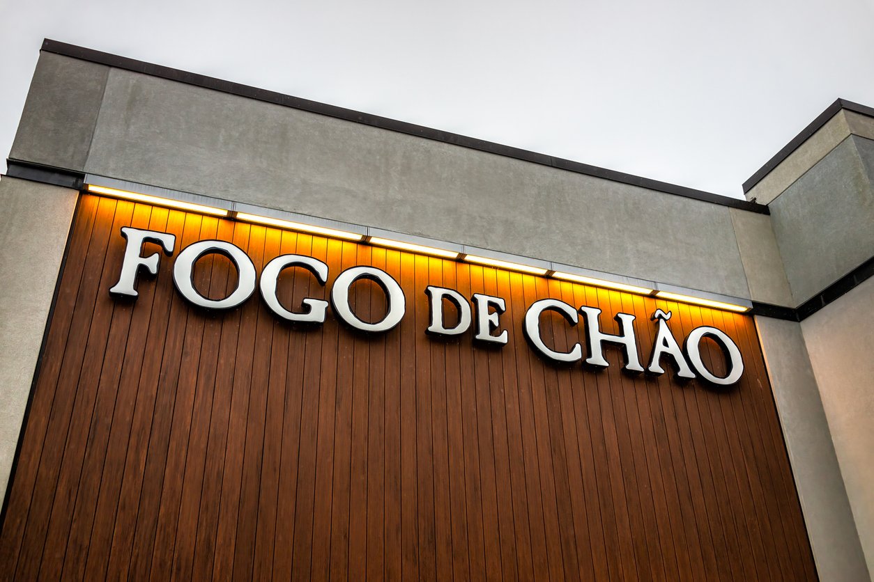 The image shows the exterior of a Fogo de Chão restaurant, featuring large, illuminated white letters with accents on a brown wooden facade beneath a gray roof—a great choice among Thanksgiving restaurants open for the holiday.