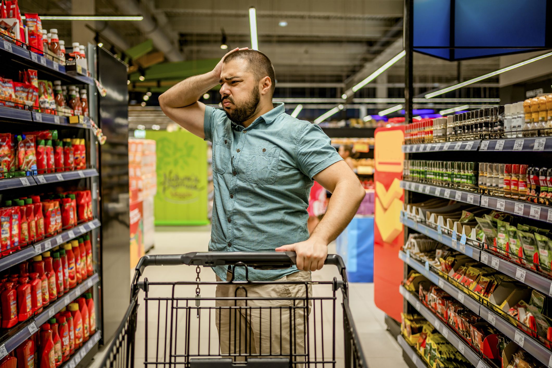 Young man standing among the produce aisle at the supermarket and feeling upset about the increase in food prices
