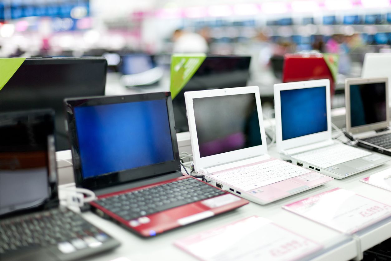 Several laptops of various colors, including black, red, white, and pink, are displayed in a row on a store counter, with a blurred background of other electronics and shelves.
