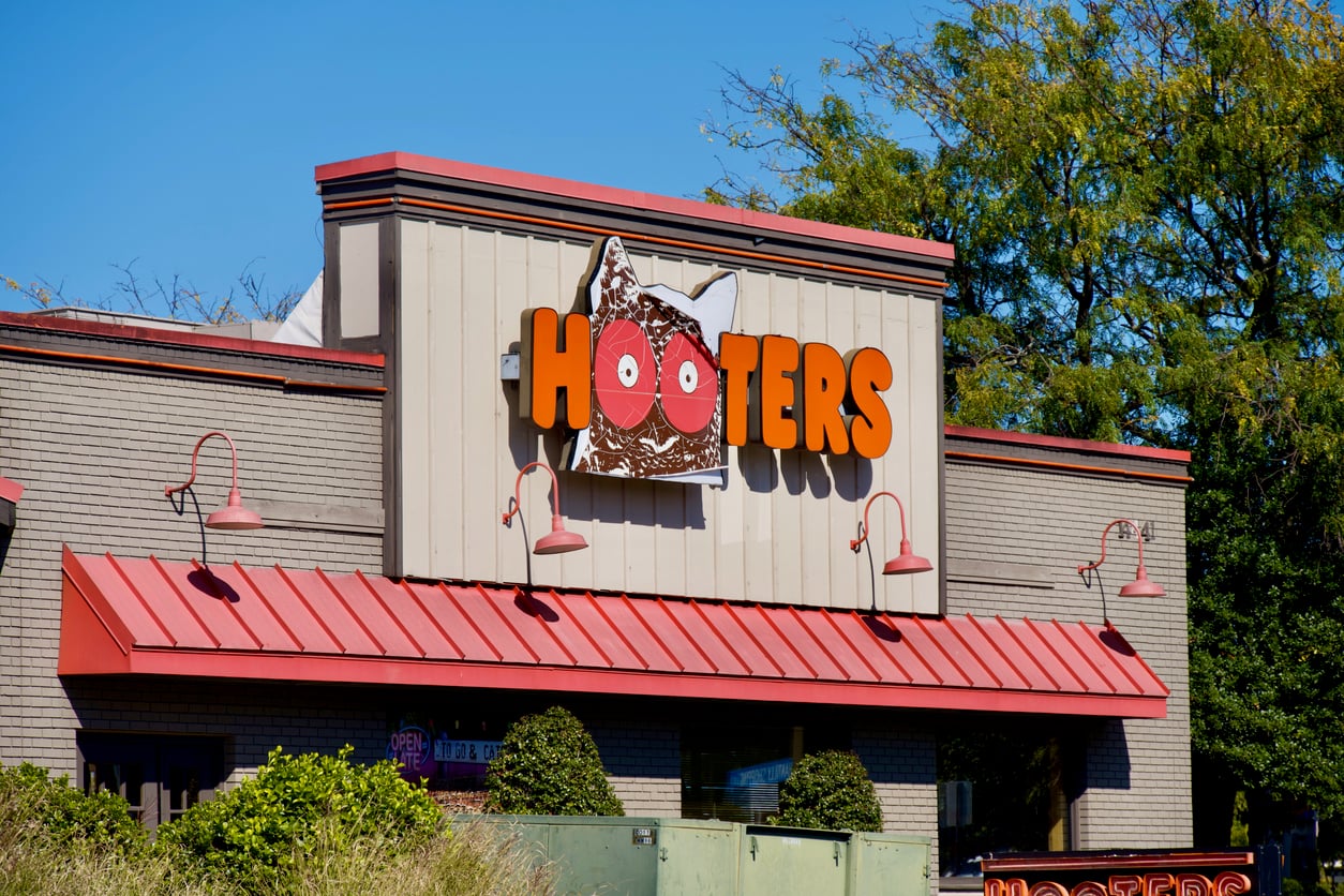 The exterior of a Hooters restaurant with a large sign featuring the restaurant’s name and an owl logo, set against a clear blue sky and surrounded by greenery—one of the Thanksgiving restaurants open for those seeking a festive meal out.