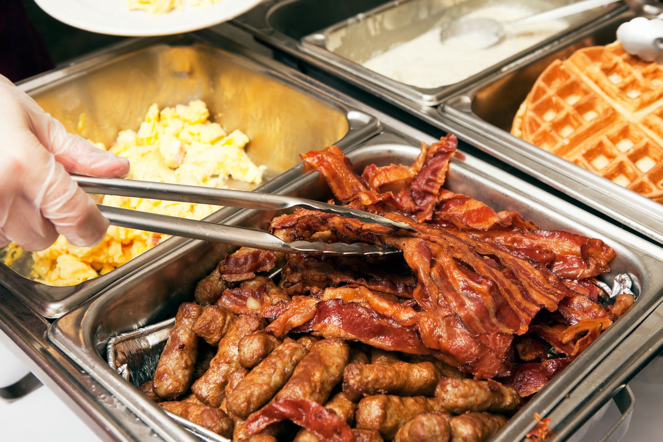 A gloved food service worker is using tongs to plate bacon from a breakfast or brunch buffet line. This chafing dish has sausage, bacon and scrambled eggs, to the right are waffles and gravy for biscuits and gravy.