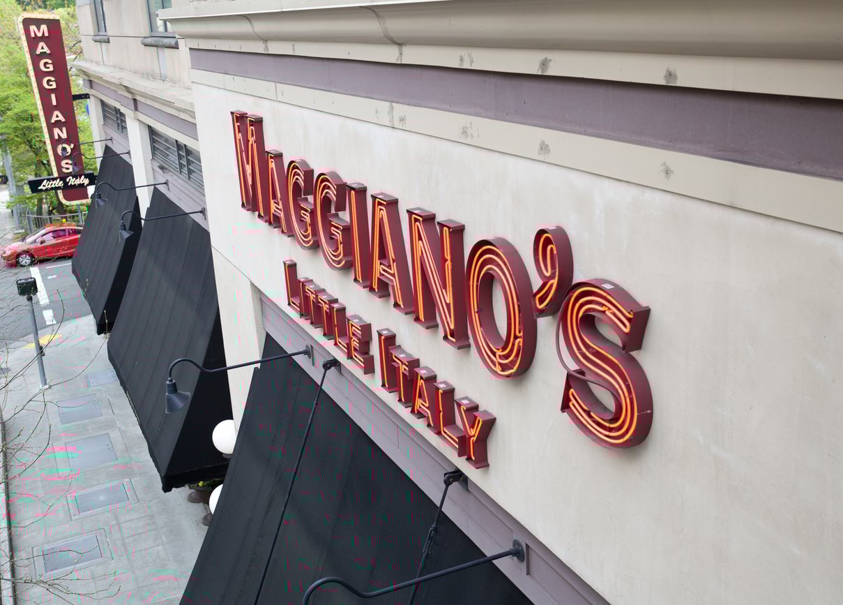 A large red neon sign for "Maggiano's Little Italy" is mounted on the side of a building above black awnings—a welcoming sight among Thanksgiving restaurants open, with a matching vertical sign visible on the street corner below.