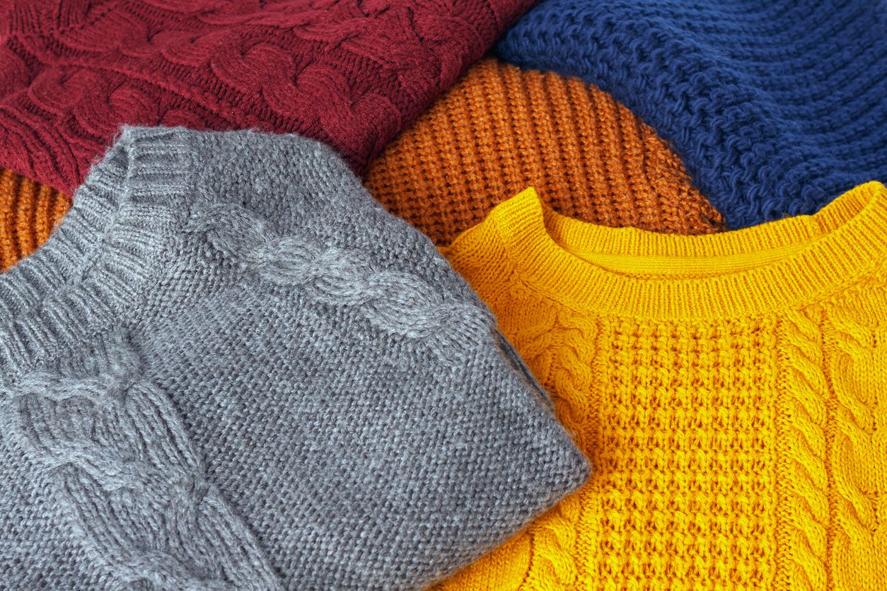 A close-up of colorful knit sweaters in red, blue, gray, yellow, and orange, arranged overlapping each other, showing detailed cable and textured patterns in the fabric.