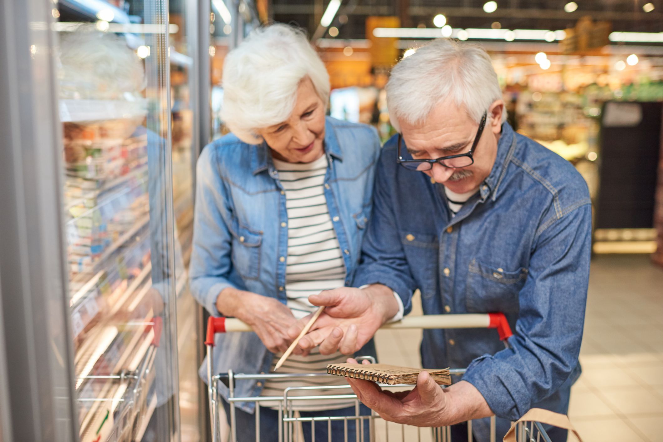Portrait of smiling senior couple grocery shopping in supermarket, reading shopping list leaning on cart standing by freezers