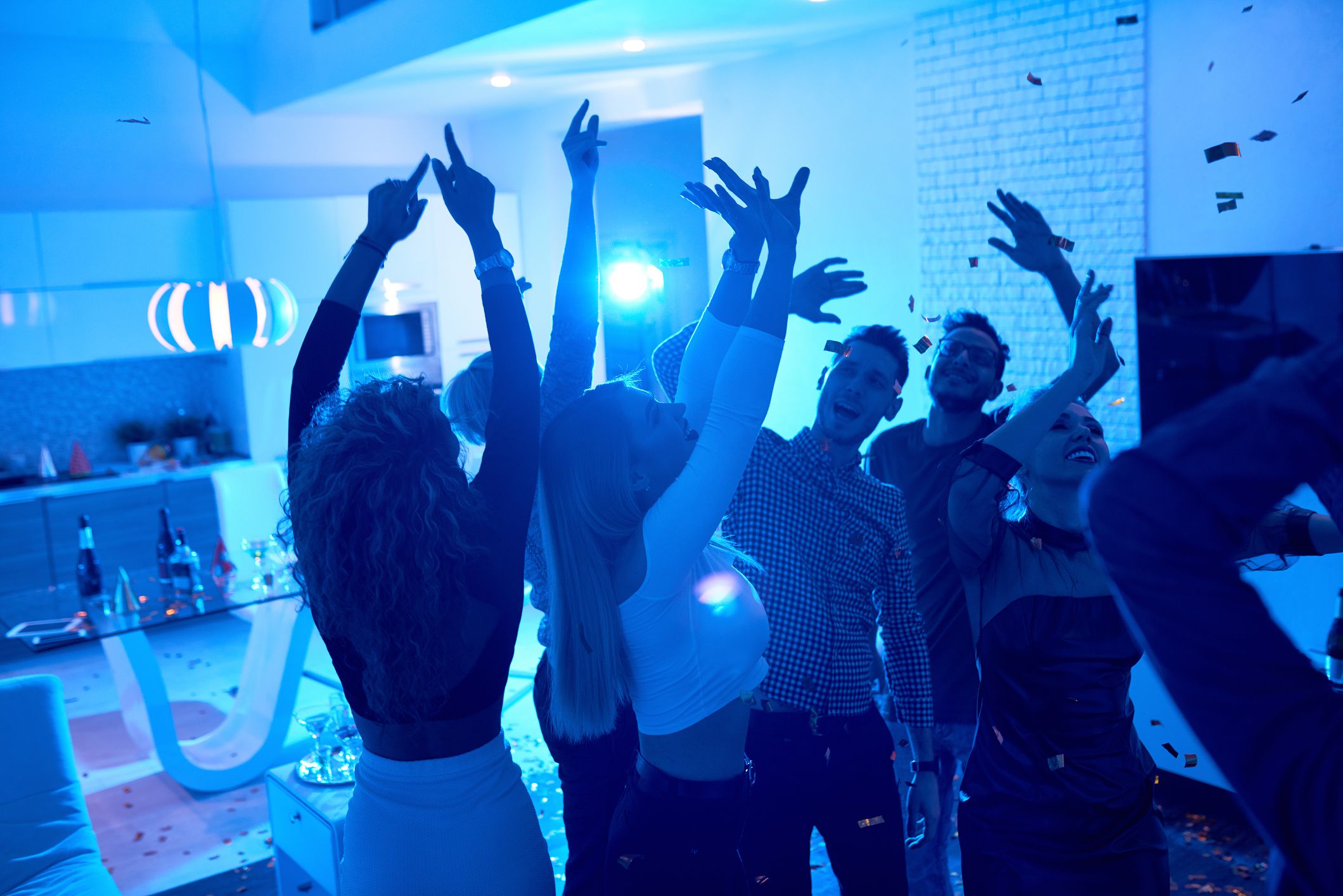 Group of modern young people dancing under confetti at private house party lit by blue light