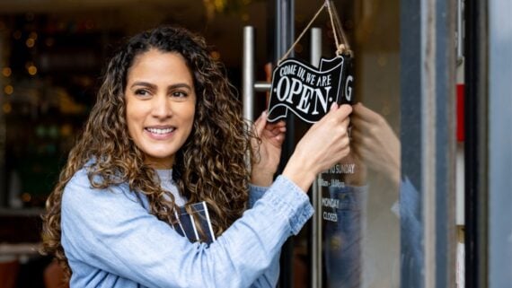 A woman with curly hair smiles as she hangs an "OPEN" sign on the door of her shop, inviting customers inside—just like Thanksgiving restaurants open to welcome guests for the holiday.