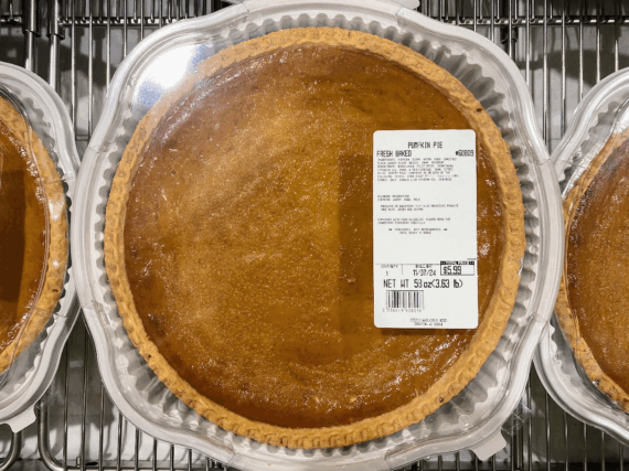 A large pumpkin pie in a clear plastic container sits on a metal shelf. A white label with product information and pricing is attached to the container’s lid. Other similar pies are visible next to it.