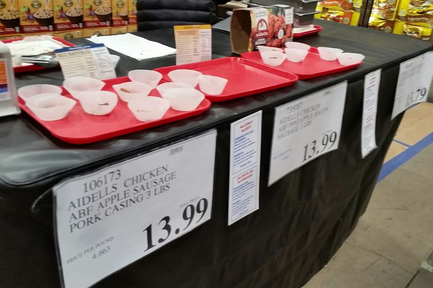 A store sampling station displays red trays with empty white sample cups on a counter. Signs below list sausage varieties and prices. Packaged goods are visible on shelves in the background.