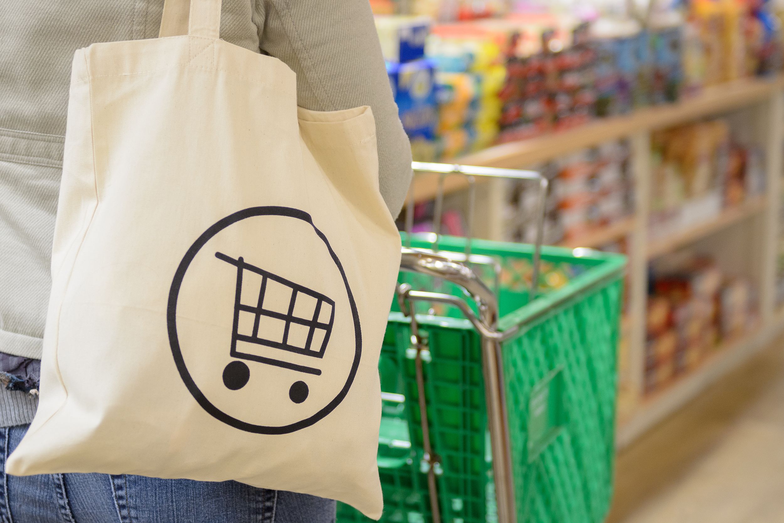 A canvas grocery bag in the foreground on the back of woman pushing a green cart, blurred background of an aisle food in a grocery store, on the right
