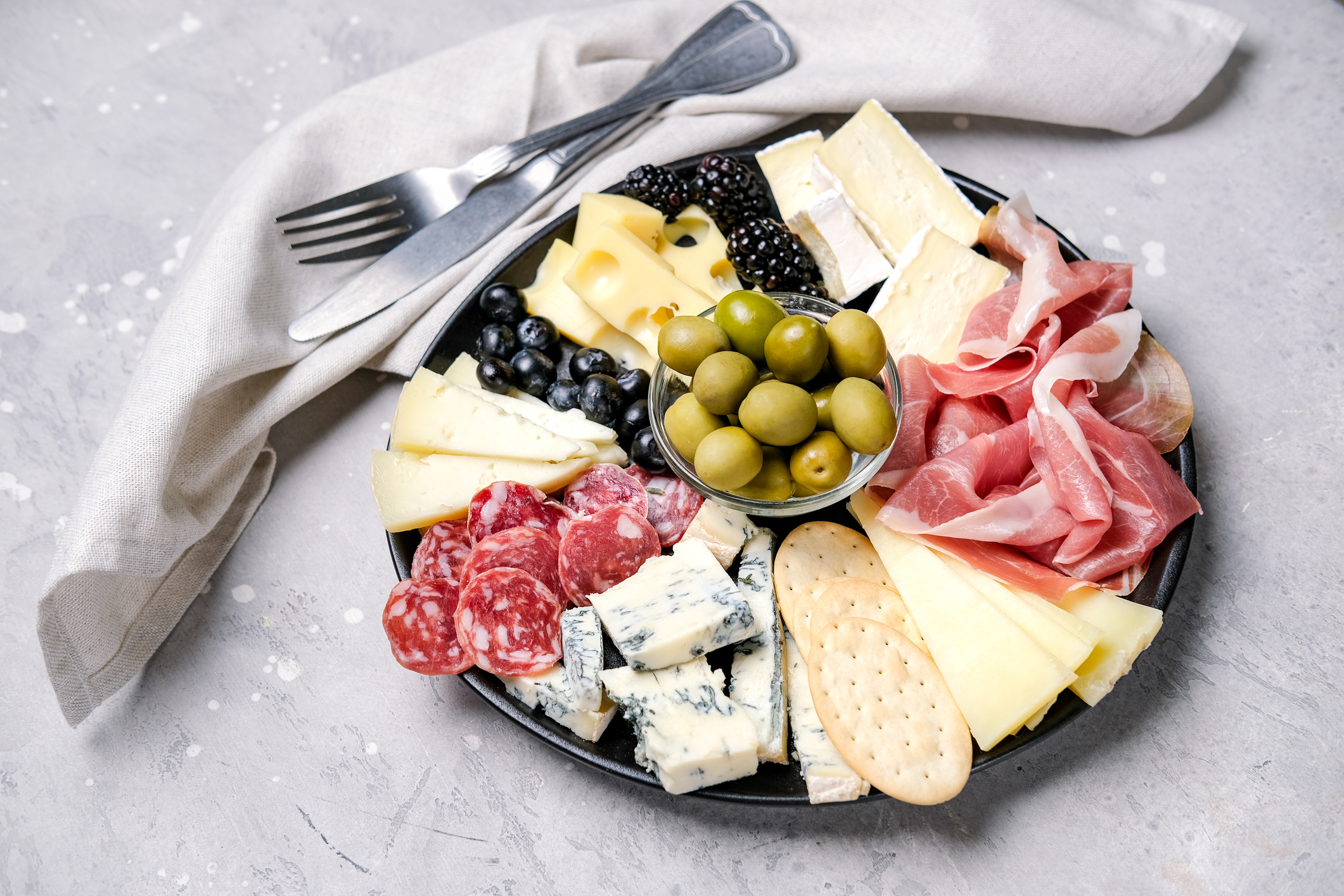 Charcuterie board with pork sausage, salami and cheese plate with brie, parmesan pecorino and gorgonzola, served with olives and blueberries, silvery grey napkin with fork and knife around it on the left to top, on a light grey table with splashes of whit