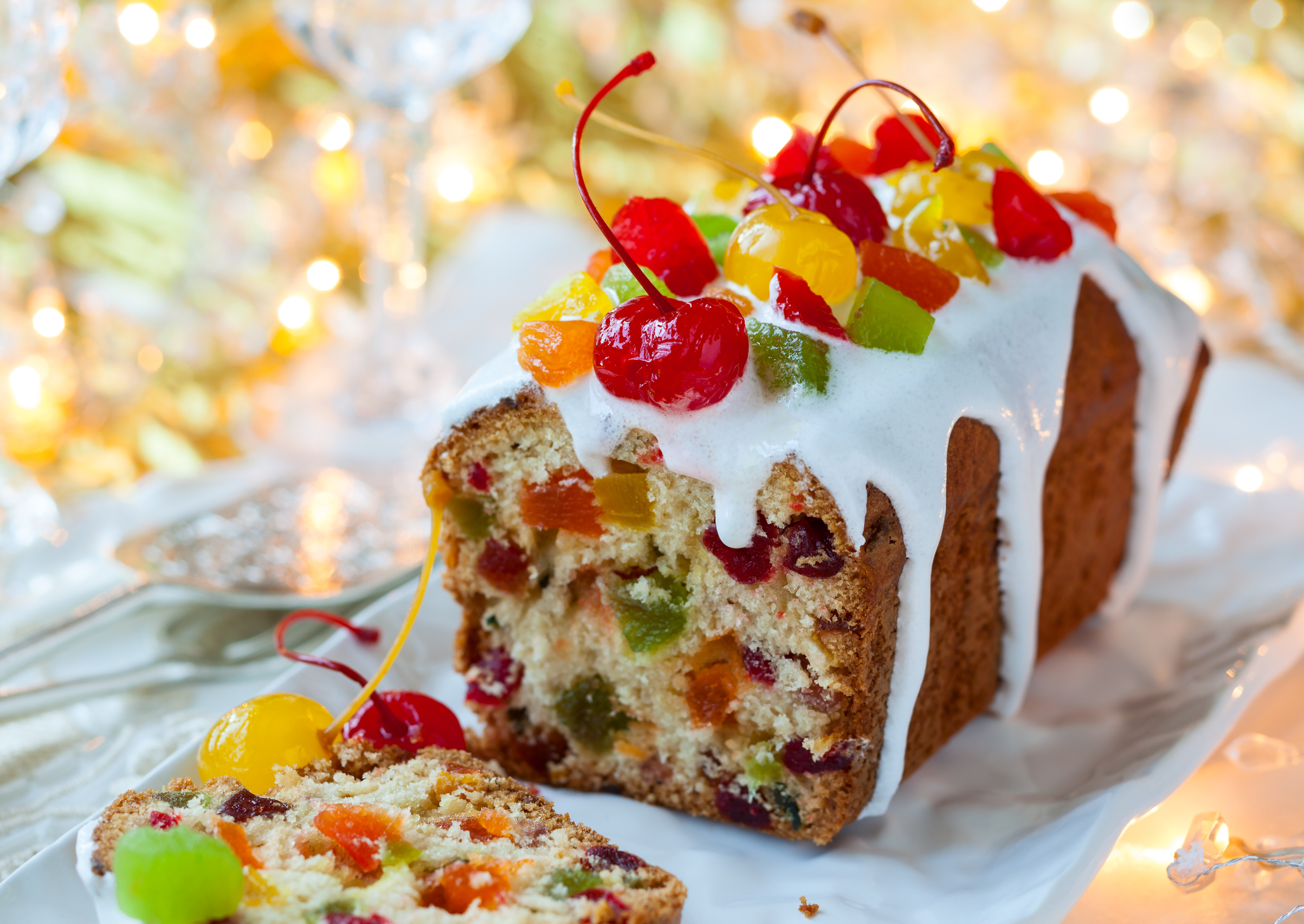 Christmas fruitcake with sugar icing and candied fruits on a white rectangle porcelain serving plate, serving silverware and white napkins to the left of it, bright Christmas tree and lights blurred in the background