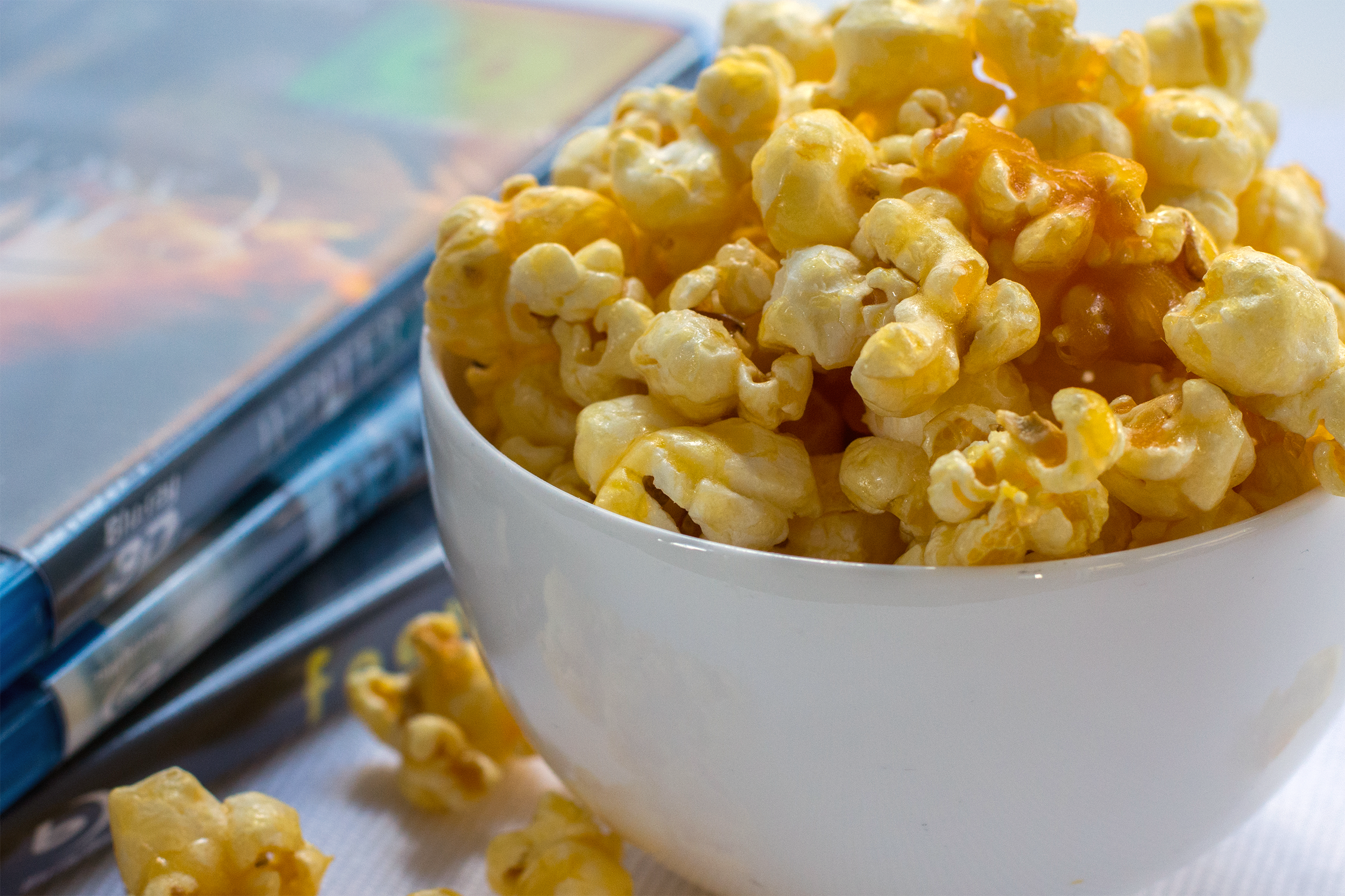 Closeup of caramel and buttered popcorn in a white porcelain bowl, on the right, three DVDs on the left, on a white napkin with pieces of popcorn