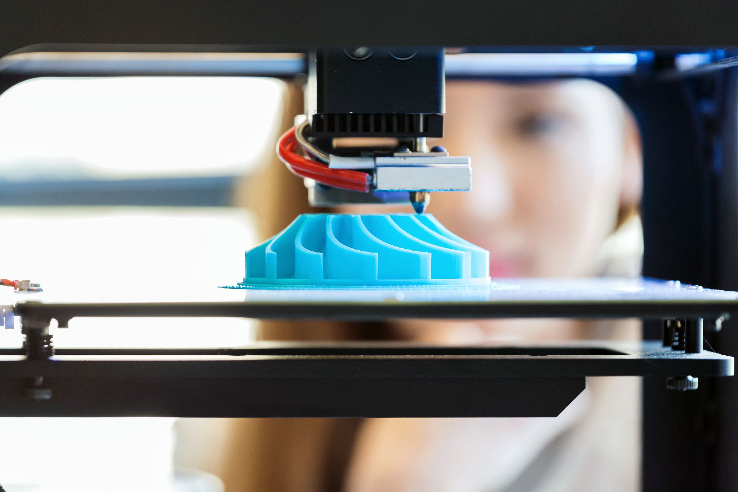 Closeup of a circular blue 3D printout being done in a 3D printer, woman watching blurred in the background