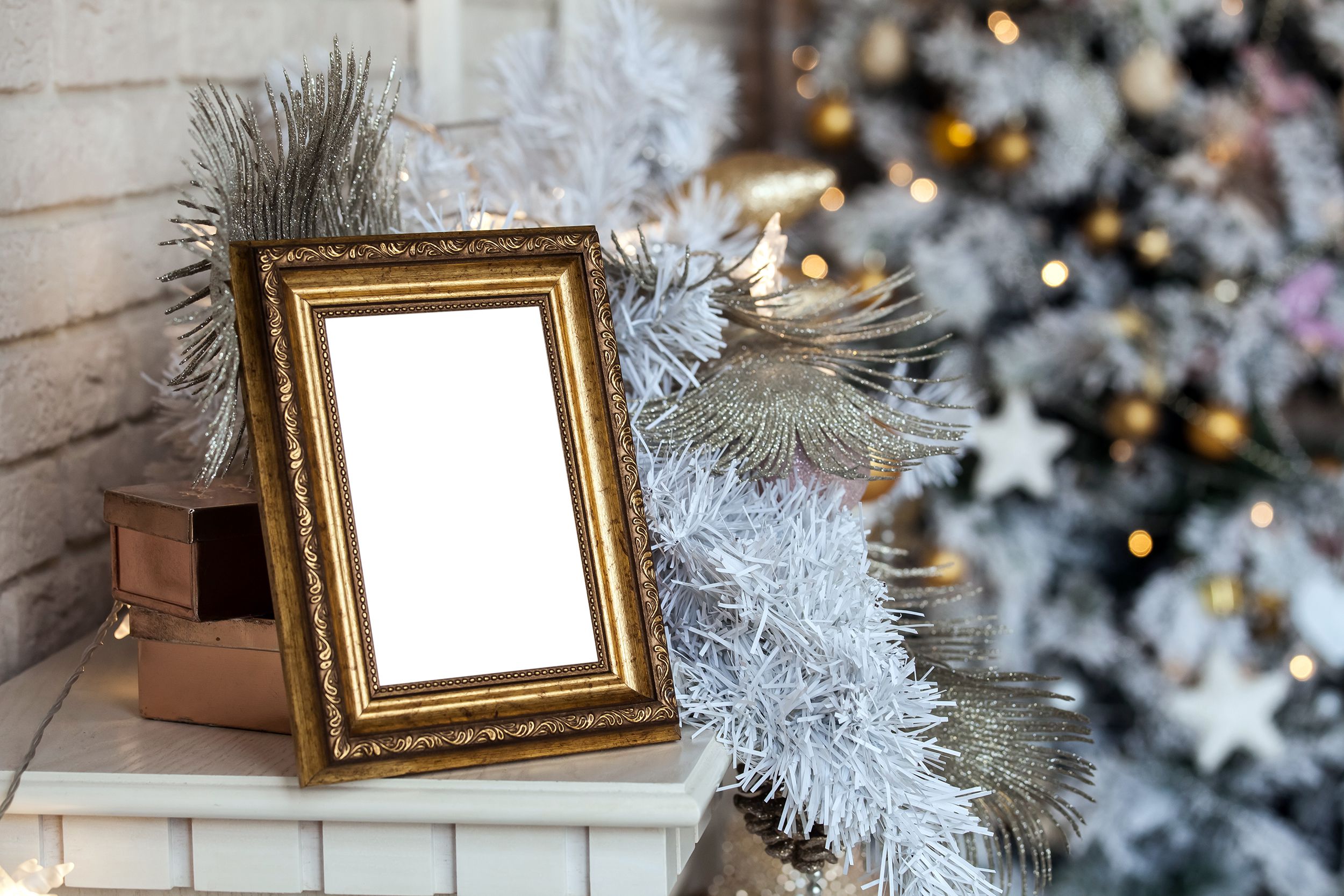 A gold ornate picture frame on a mantle with silvery and snowy holiday decorations, in the foreground left, a decorated Christmas tree blurred in the background right