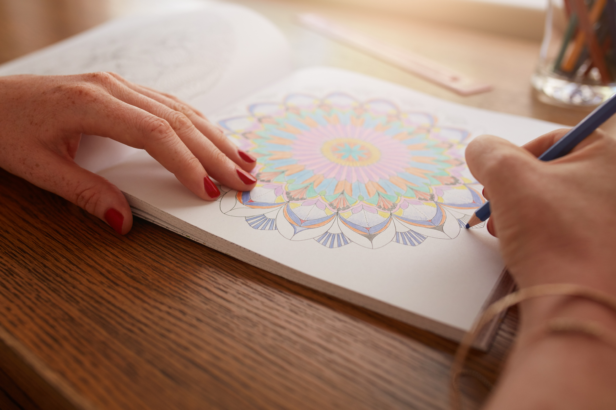 Closeup of female hands coloring with a blue colored pencil in an adult coloring book at a wooden desk, selective focus, colored pencils in a glass and a ruler on the top right, blurred in the background