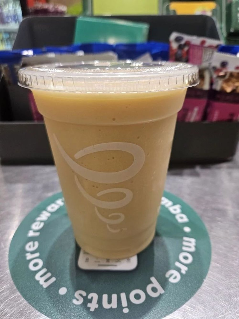 A plastic cup filled with a creamy, light yellow smoothie—reminiscent of a fast food secret menu item—sits on a circular green sign that reads "more rewards. more points." The background features packaged snacks and part of a checkout counter.
