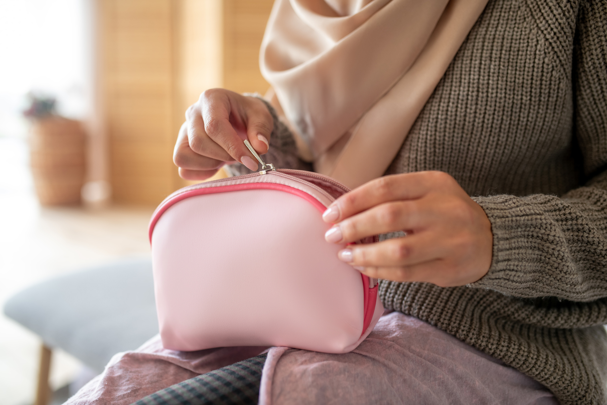 Light pink cosmetics bag being zipped by two hands of a woman, on her lap, selective focus, blurred background of light blue bench she is sitting on, white floor, light brown walls, and sunlight on the left