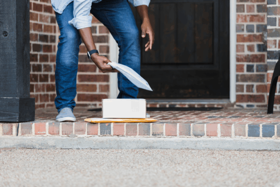 Man slightly squatting, picking up a package outside his front door, package is on a pile of two other packages, porch is brick, shows legs, arms, and midsection of man, concrete slab in the foreground