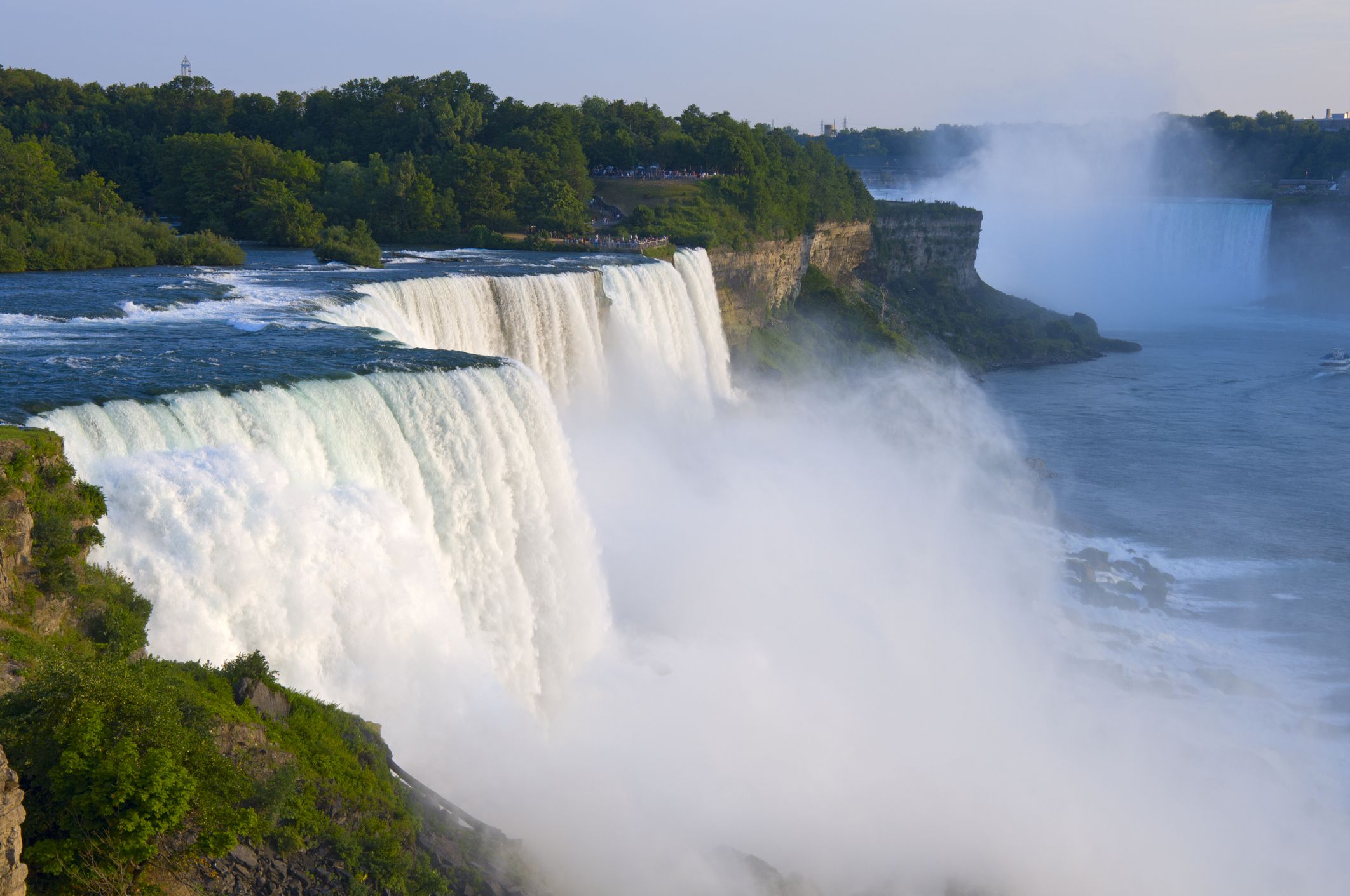 A wide view of Niagara Falls shows powerful waterfalls cascading over cliffs, creating mist that rises above the river, with lush green trees in the background under a clear sky.