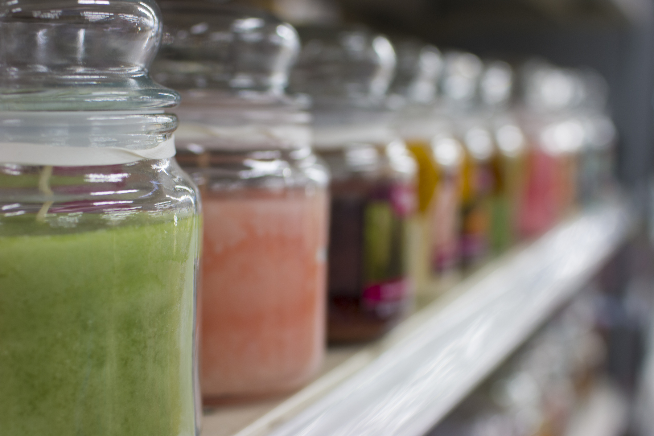 Row of glass jar candles, different colors, green candle on the left foreground, selective focus, on a white shelf at a store, blurred bottom right