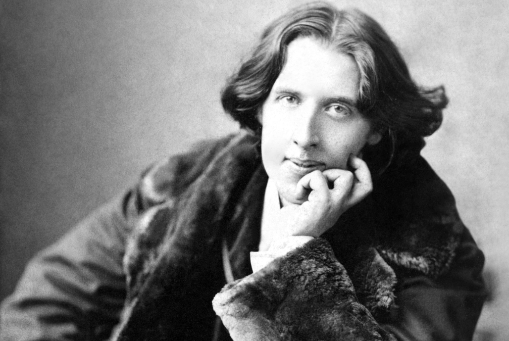 Black-and-white portrait of a person with wavy hair, wearing a fur-collared coat, resting their chin on their hand and gazing thoughtfully at the camera. The background is plain and softly blurred.