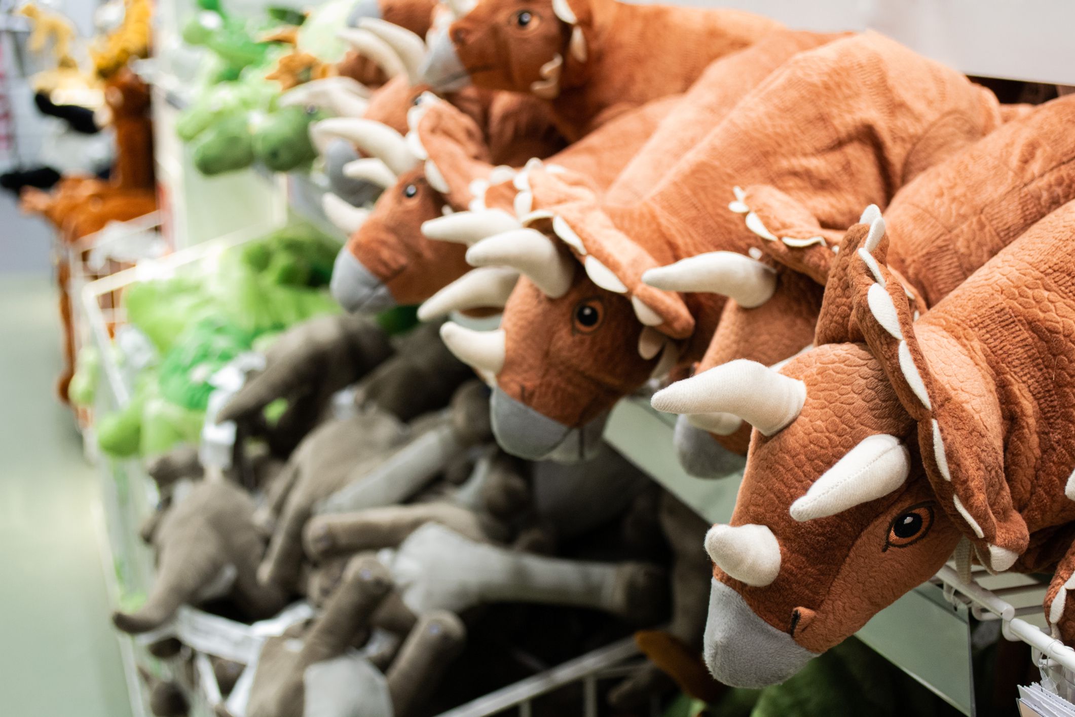Selection of stuff animal dinosaurs for sale, selective focus of brown triceratops, two rows of brown, grey, and green, different kinds of dinosaurs, blurred with a grey floor at a store