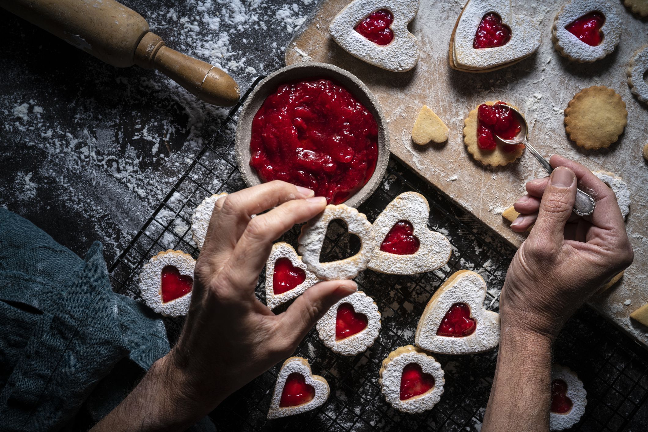Top view of two hands making almond & jam cookies, one hand putting jam on a cookie, surrounded by flour, baking ingredients, and rolling pin