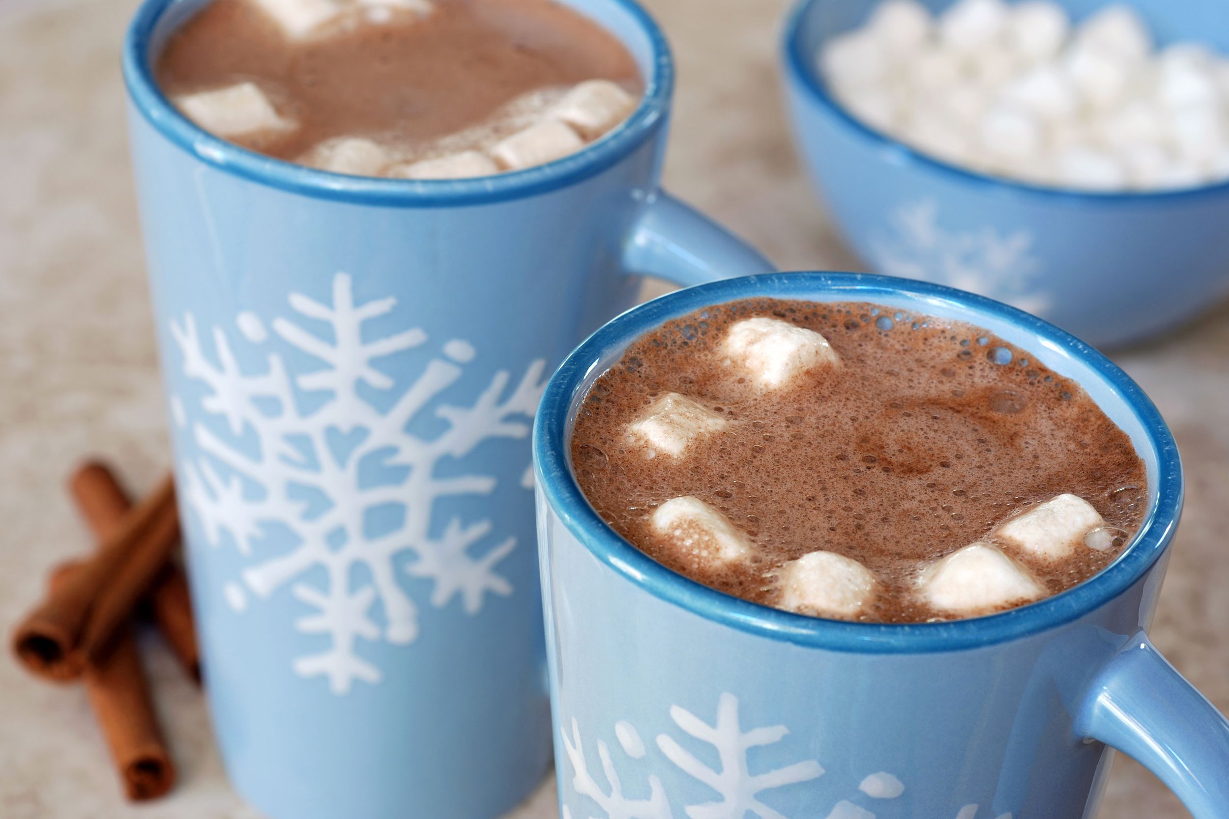 Top-view of two long light blue with a white snowflake design mugs filled with hot chocolate and small marshmallows, selective focus, blurred background of three cinnamon sticks by the left one and a matching light blue bowl of marshmallows on the top