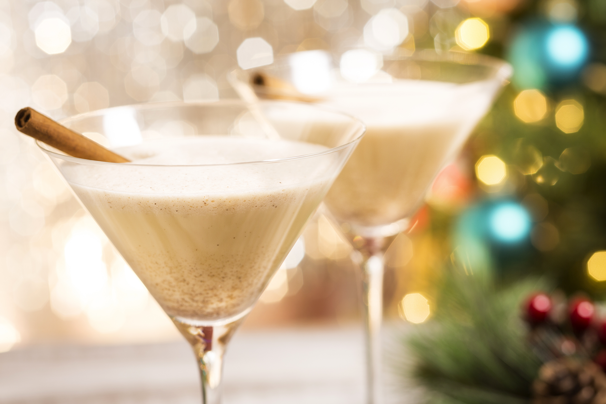 Two martini glasses with spiked eggnog, left one is selective focus, right one is somewhat blurred, a cinnamon stick hangs out in each, blurred sparkles on the background left with Christmas tree with lights on the background right