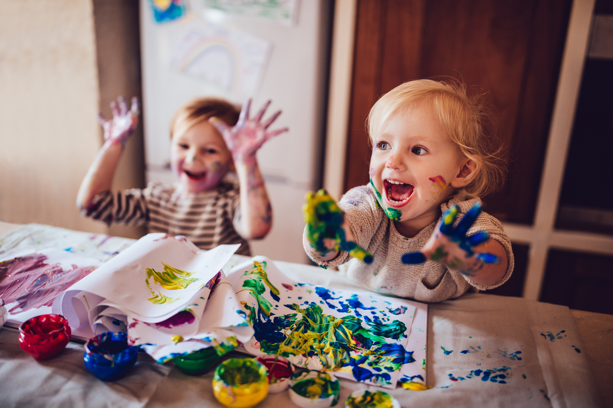 Two toddlers playing with finger paints, both are smiling and laughing, male on the right in selective focus, female blurred on the left, artwork, paper, and paints on the table in the foreground, beige wall and refrigerator blurred in the background