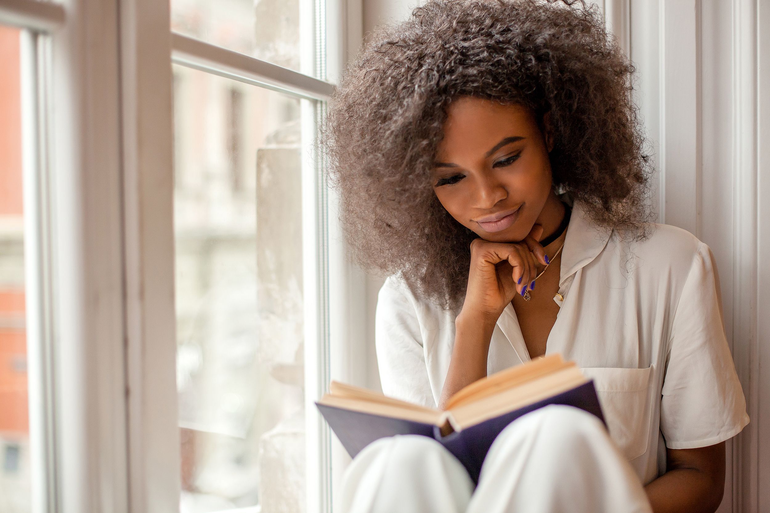 Woman reading a book while sitting on a window sill, on the right, window of building in a city, on the left