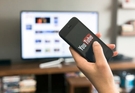 A hand holds a smartphone displaying the YouTube logo, with a TV screen showing a blurred interface in the background, suggesting video streaming or casting between devices&mdash;perfect for exploring subscriptions worth the money.