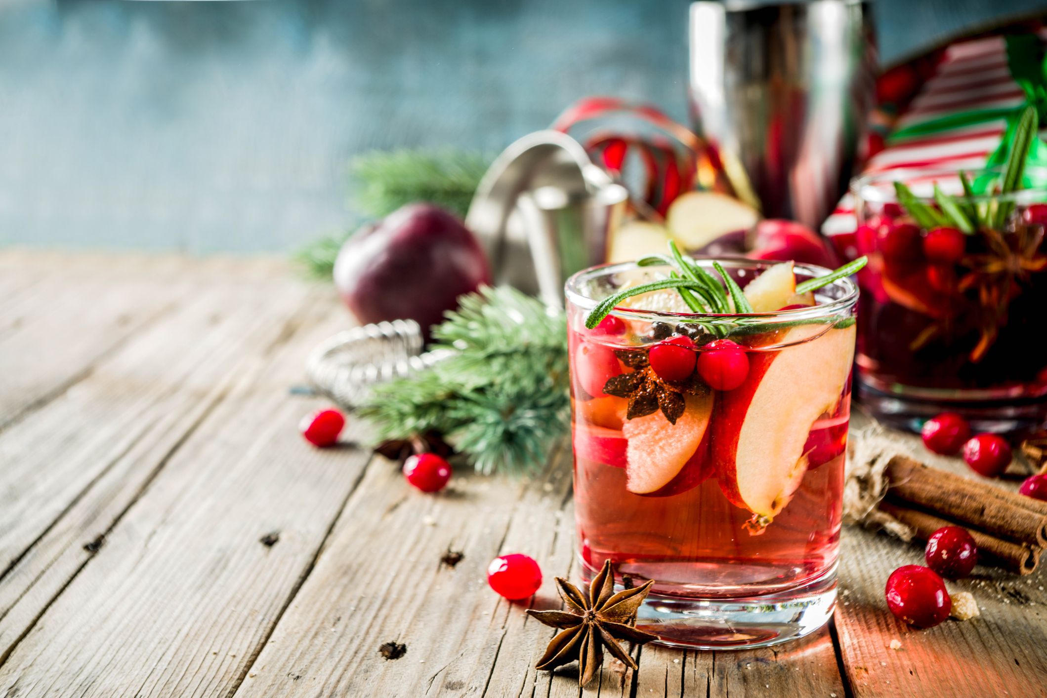 A festive drink garnished with apple slices, cranberries, rosemary, and star anise sits on a wooden table, surrounded by cranberries, cinnamon sticks, pine branches, and holiday decorations.