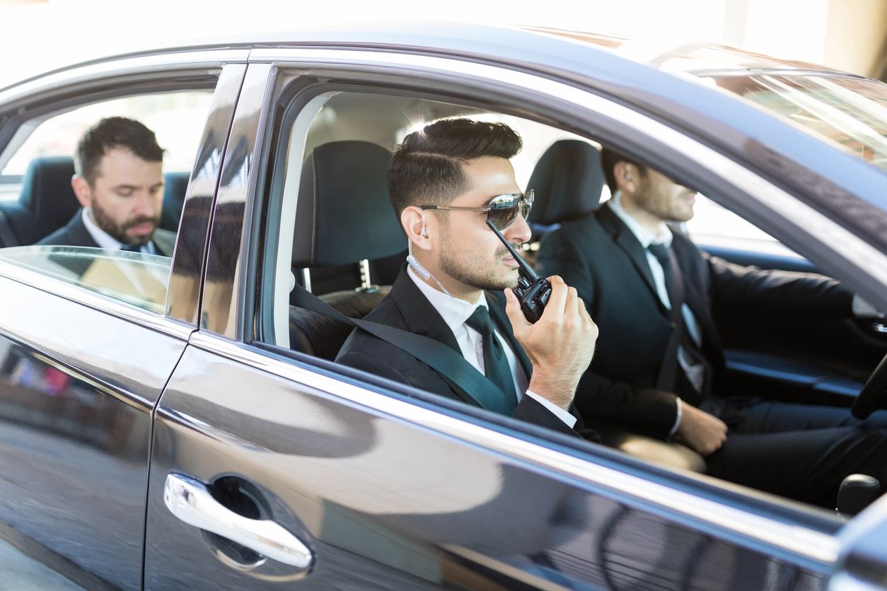 A businessman who is sitting in the back of a nice car while being transported by two security guards.