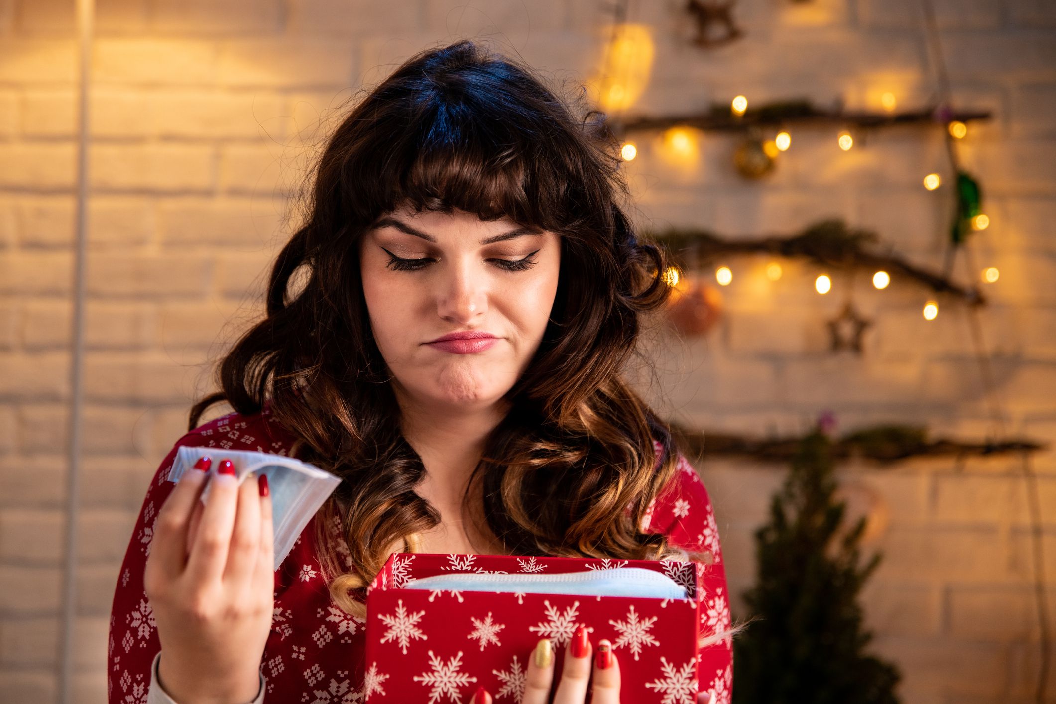 Lousy awful Christmas gift. Lovely woman in winter pajamas opening Christmas gift box with surgical face masks. She sits in the living room with the alternative Christmas tree made of branches in the background on a white brick wall.