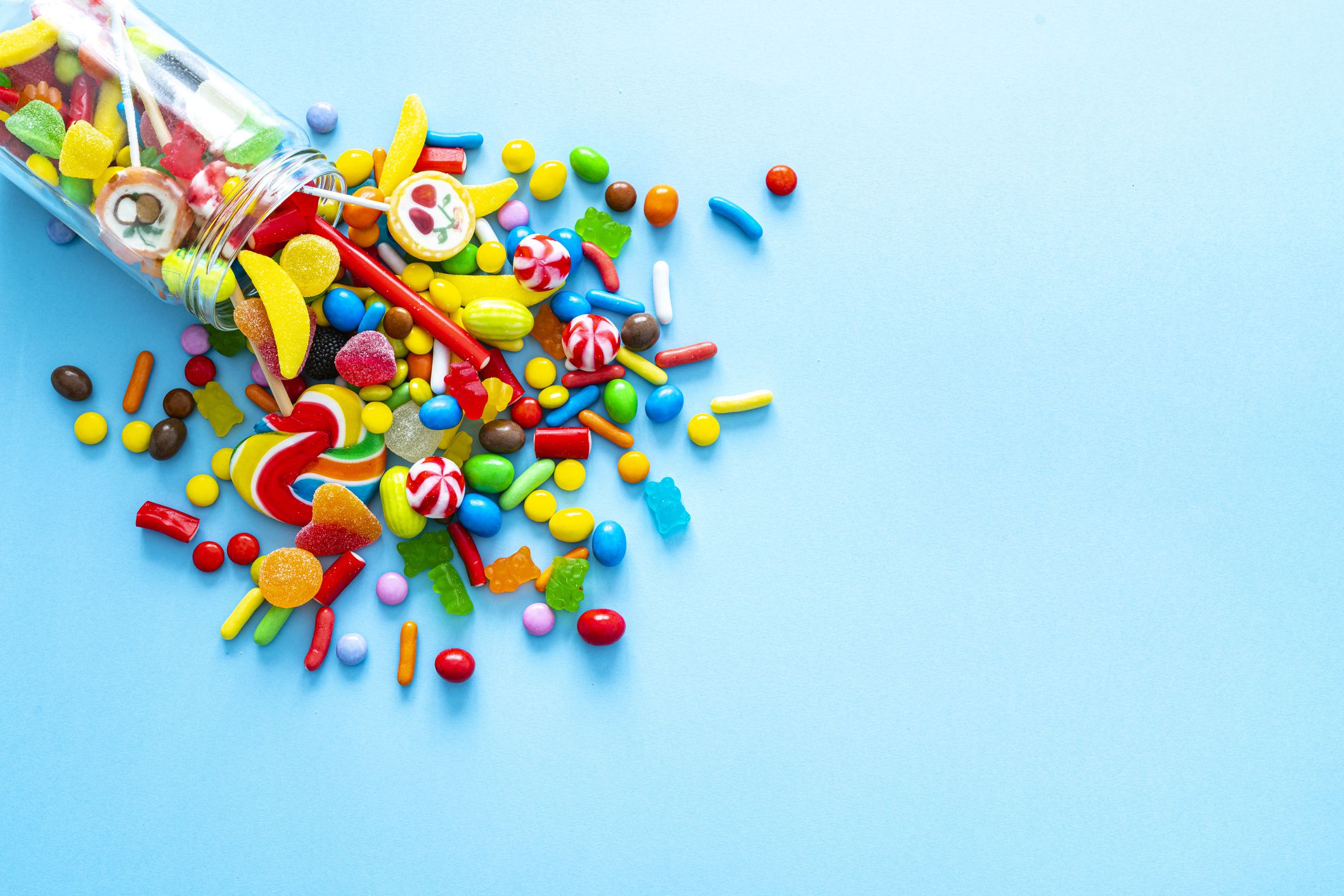 Large group of all sort of multicolored candies spilling out from a glass jar shot from above on blue background. High resolution 42Mp studio digital capture taken with SONY A7rII and Zeiss Batis 40mm F2.0 CF lens