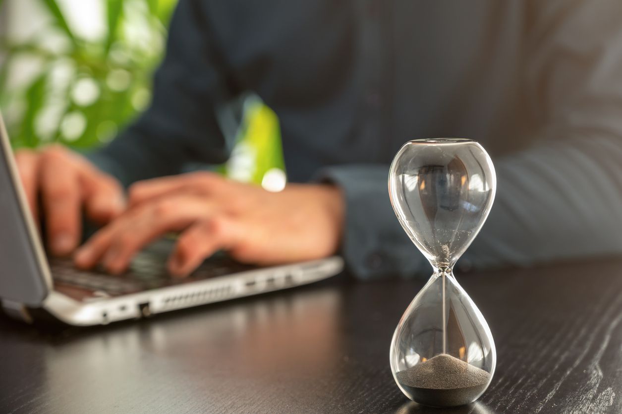 A person typing on their laptop with a sand timer placed next to it.