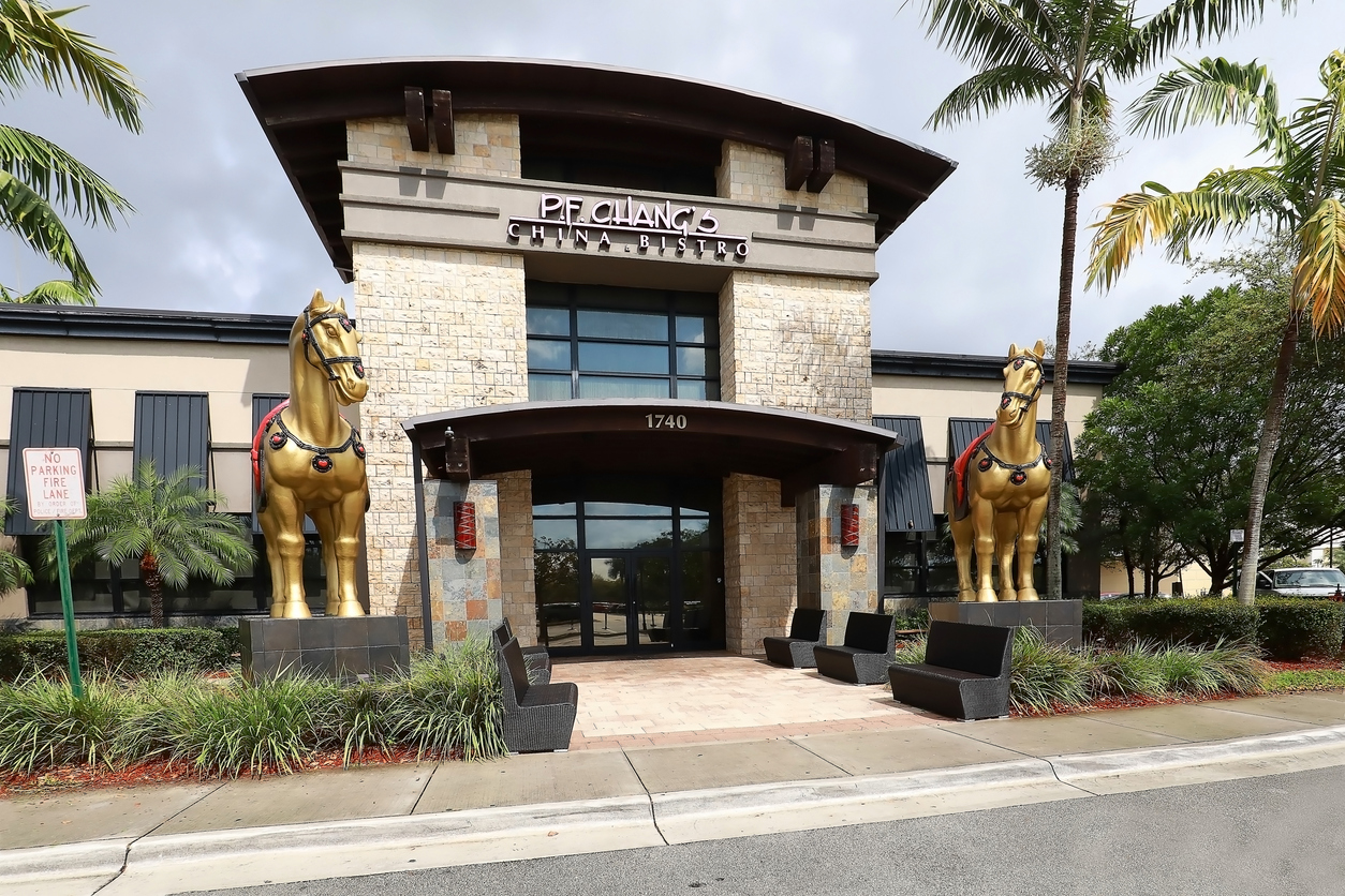 The entrance of P.F. Chang's China Bistro features two large golden horse statues on either side, with palm trees and modern stone architecture under a cloudy sky.