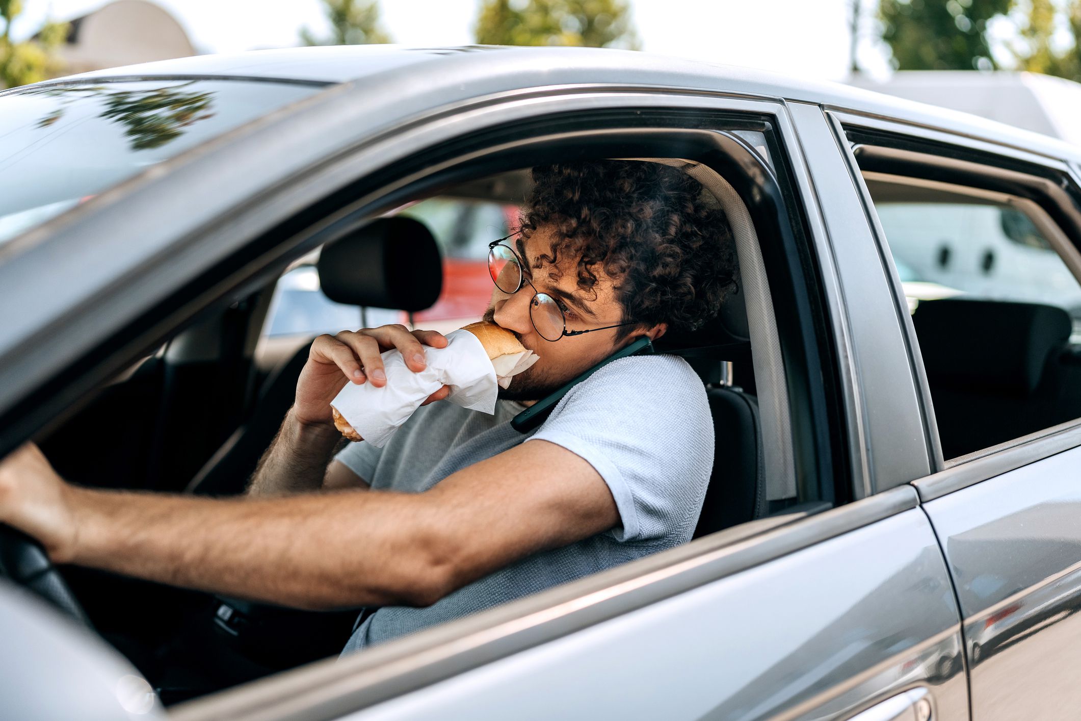 Young casually clothed man talking on a phone and eating sandwich while driving car