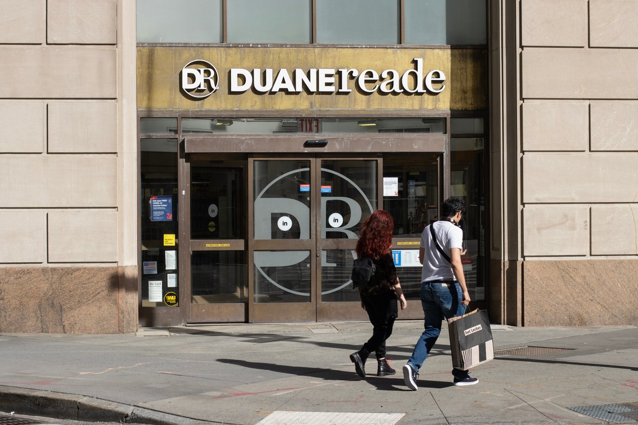 Two people walk past the entrance of a Duane Reade store. The storefront features a large sign above the door and a smiley face design on the glass doors. One person carries a shopping bag.