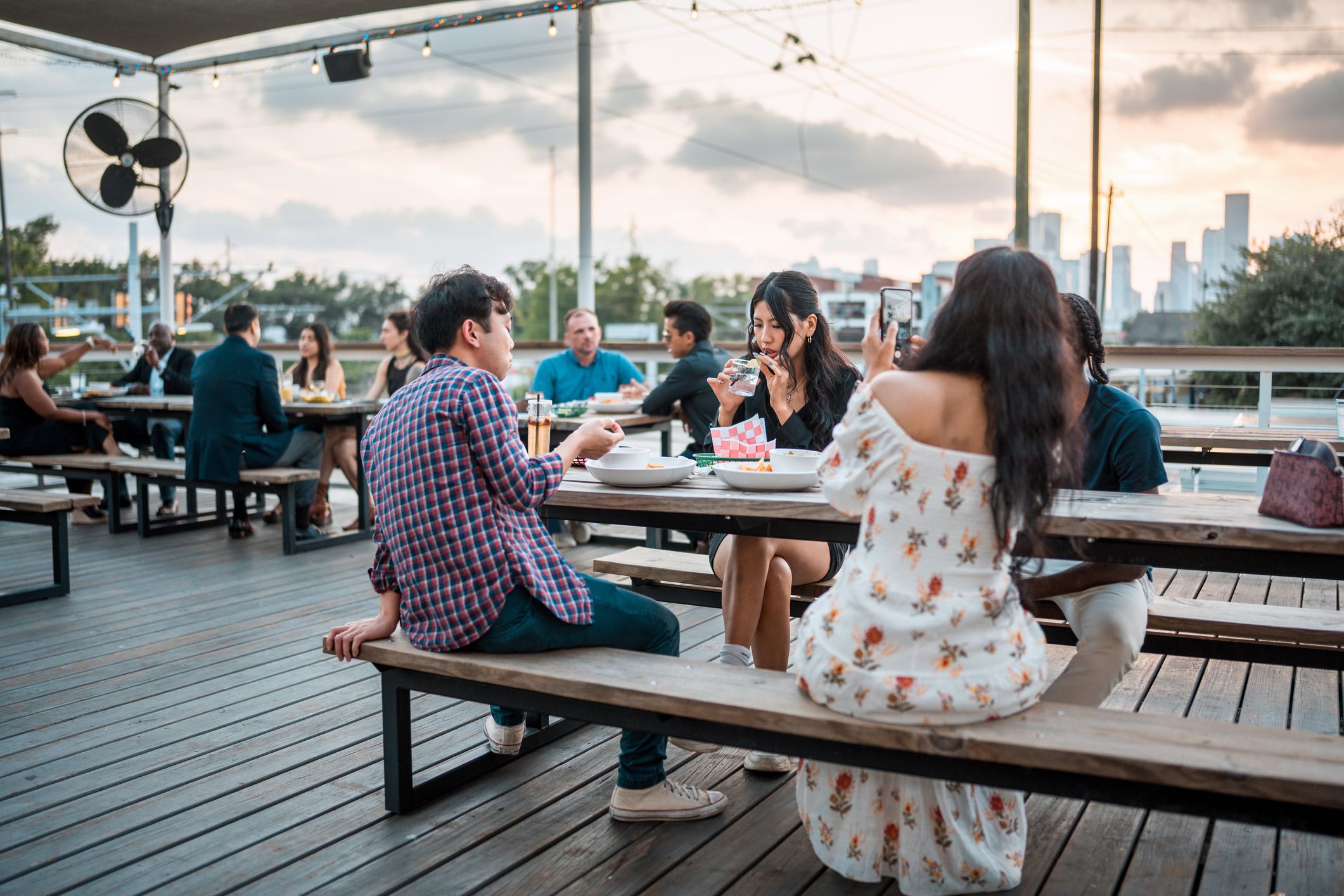 A cheerful diverse group relishes an outdoor meal together on a bustling restaurant terrace, surrounded by the gentle dusk light.