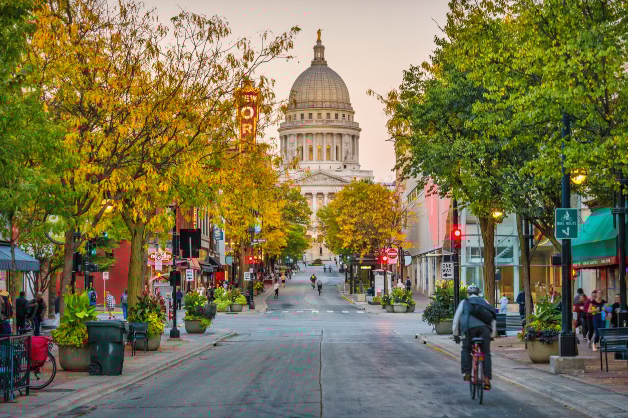 A wide street lined with trees and shops leads to a large domed capitol building. People are walking and riding bikes, and trees display early autumn colors under a soft, warm sky.