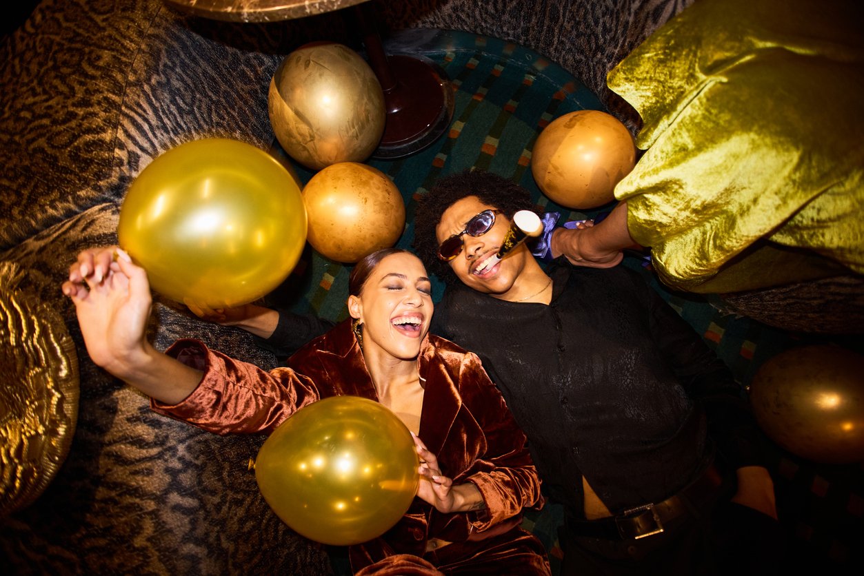 Two people lie on the floor, smiling and laughing, holding gold balloons at a festive party. They are surrounded by more balloons and colorful decor, creating a joyful and celebratory atmosphere.