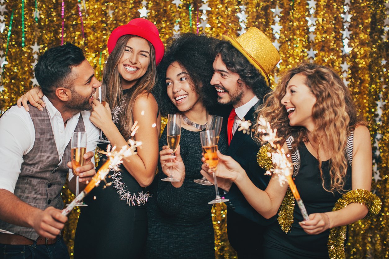 Five friends celebrate at a party, smiling and holding sparklers and champagne glasses. They wear festive hats and stand in front of a gold tinsel backdrop decorated with stars.